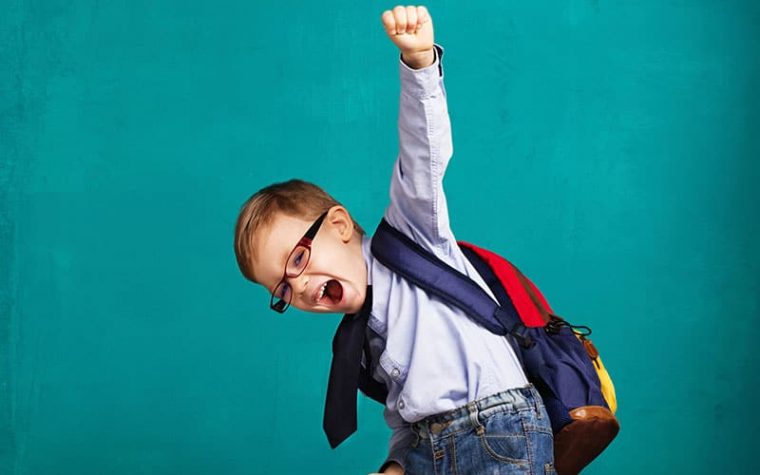 Cheerful smiling little boy with big backpack jumping and having fun against blue wall. Looking at camera. School concept. Back to School