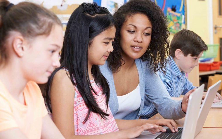 Teacher Helping Group Of Elementary School Children In Computer Class