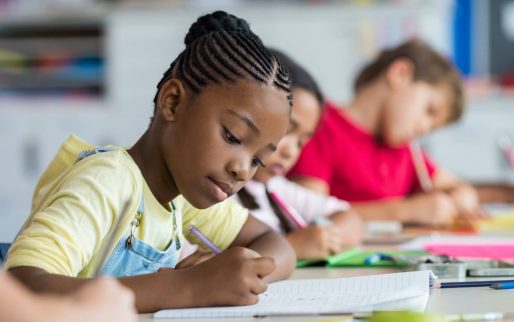 Cute pupil writing at desk in classroom at the elementary school. Student girl doing test in primary school. Children writing notes in classroom. African schoolgirl writing on notebook during the lesson.