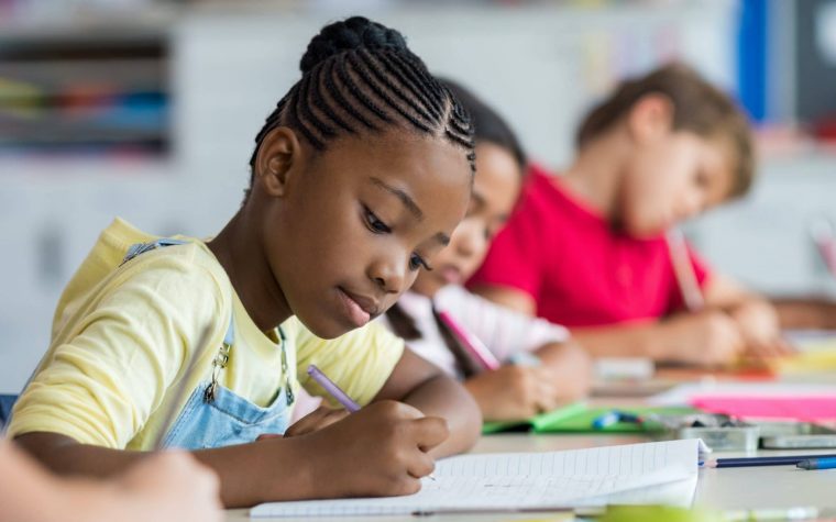 Cute pupil writing at desk in classroom at the elementary school. Student girl doing test in primary school. Children writing notes in classroom. African schoolgirl writing on notebook during the lesson.