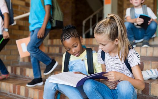 Two young girls sitting together on stairs and studying after school. Elementary schoolgirls studying the notes on copybook. Happy african girl and her friend doing homework on notebook at primary school.