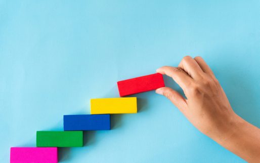 Concept of building success foundation. Women hand put red wooden block on colorful wooden blocks in the shape of a staircase