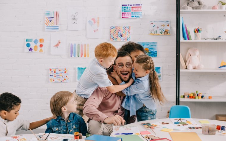 interracial kids hugging happy teacher at table in classroom