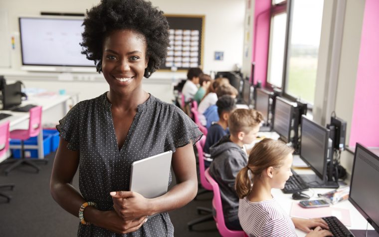 Portrait Of Female Teacher Holding Digital Tablet Teaching Line Of High School Students Sitting By Screens In Computer Class