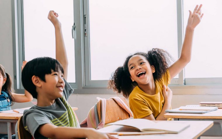 Elementary age Asian student boy raised hands up in class. Diverse group of pre-school pupils in elementary age in education building school. Volunteering and participating classroom concept.