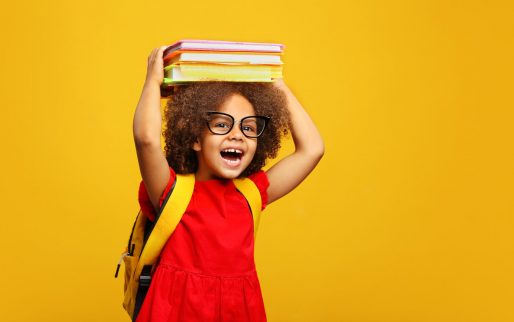 funny smiling Black child school girl with glasses hold books on her head. Yellow background