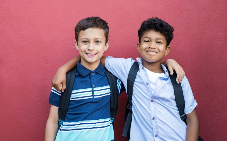 Best children friends standing with hand on shoulder against red background. Happy smiling classmates standing together on red wall after school. Portrait of multiethnic schoolboys enjoying friendship.