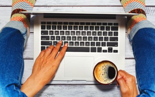 Young female person dressed in blue jeans and cozy knitted socks using a laptop by typing on keyboard and drinking coffee while sitting on the wooden floor in loft style. First person high angle view.