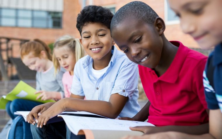 Multiethnic children sitting in a row and reading from notebook all together. School kid revising notes for exams sitting on the steps outside the elementary school. Latin school boy studying with friends before classwork.