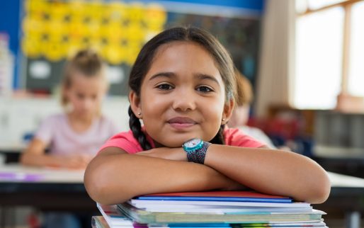 Portrait of cute little schoolgirl leaning on stacked books in classroom. Happy young latin girl in casual keeping chin on notebooks. Closeup face of smiling girl at elementary school.