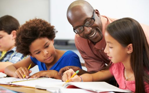 Teacher Helping Pupils Studying At Desks In Classroom Checking Work Smiling